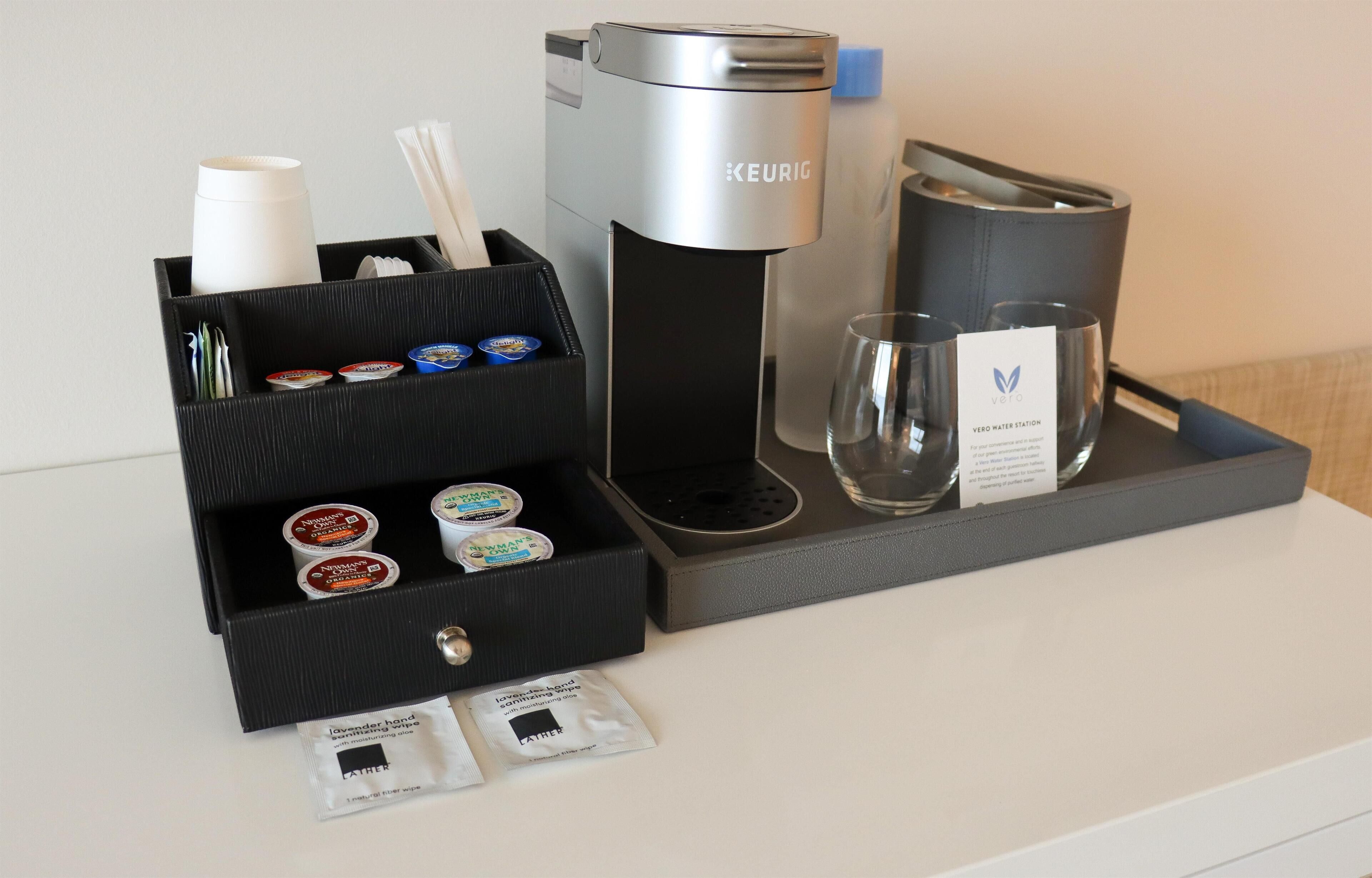 En kaffestation med en Keurig-kaffemaskin, engångsmuggar, ett glas och kaffekapslar.