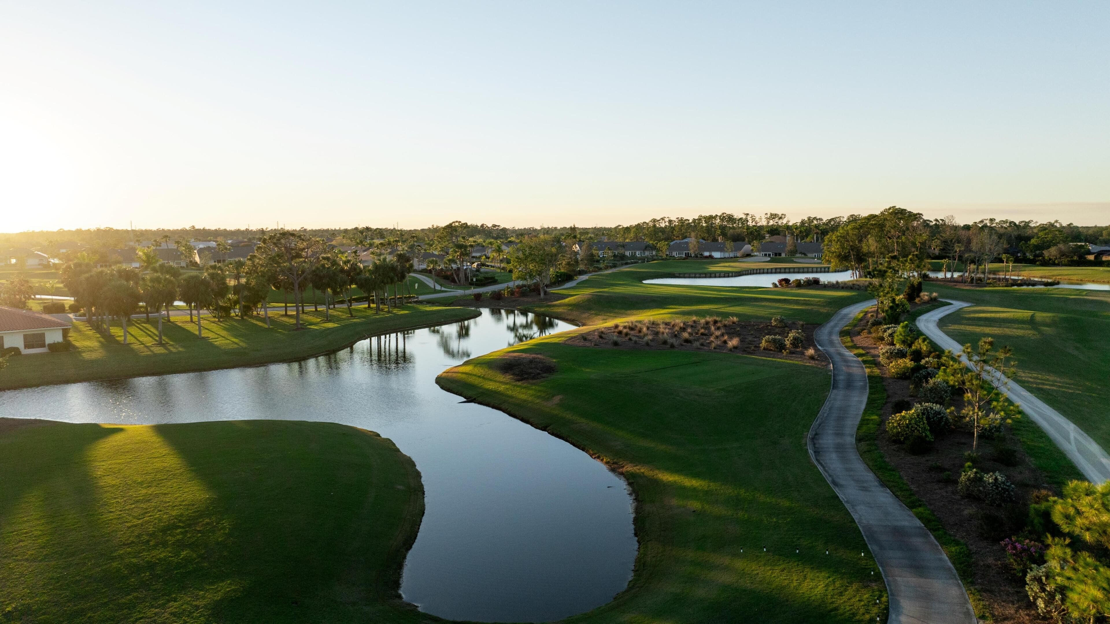 Um campo de golfe com um lago, caminhos e casas residenciais ao fundo.