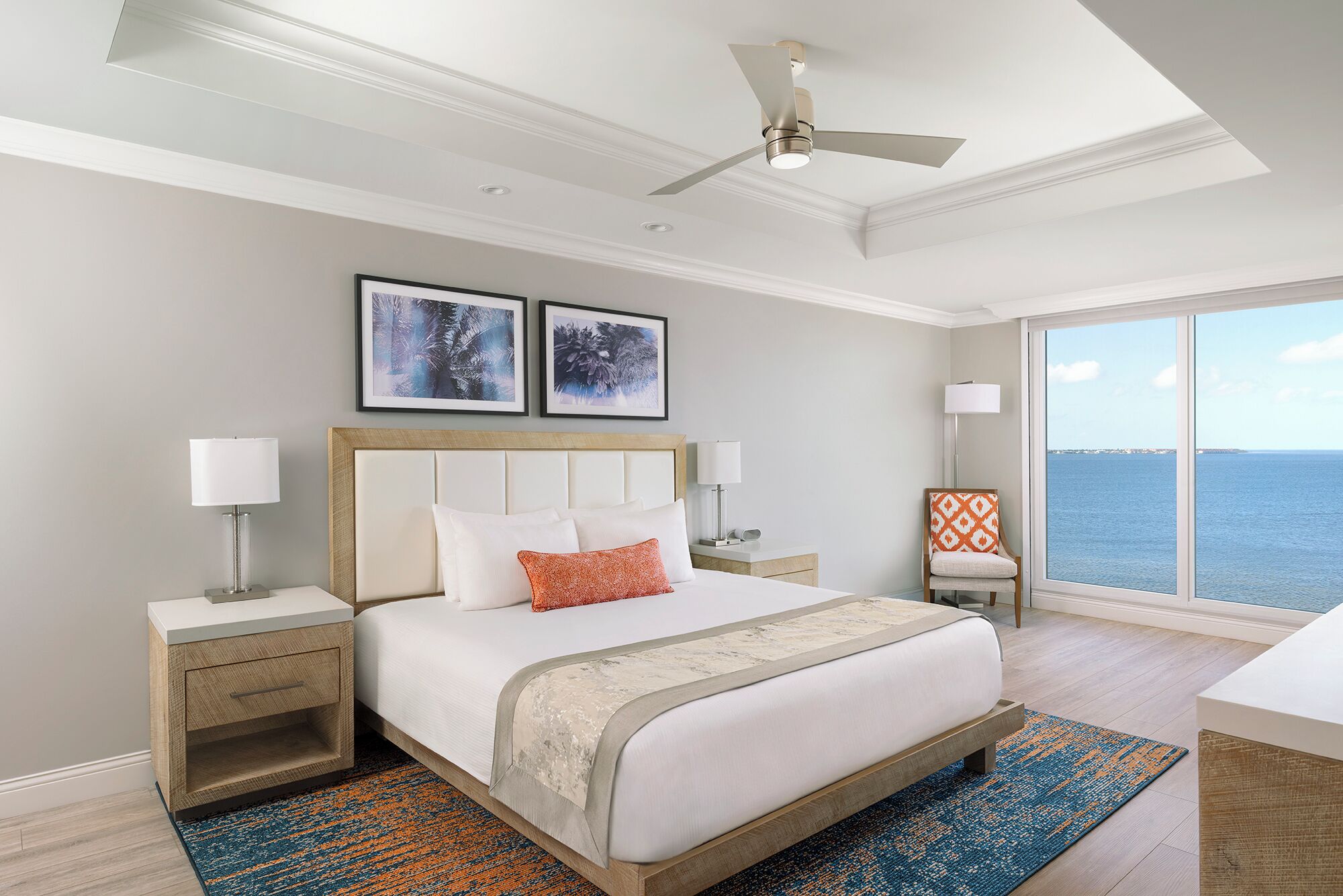 A hotel room with a bed, nightstands, a chair, a large window with a view, and ceiling fans.