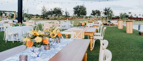 An outdoor event space with tables set for guests, surrounded by greenery and string lights.