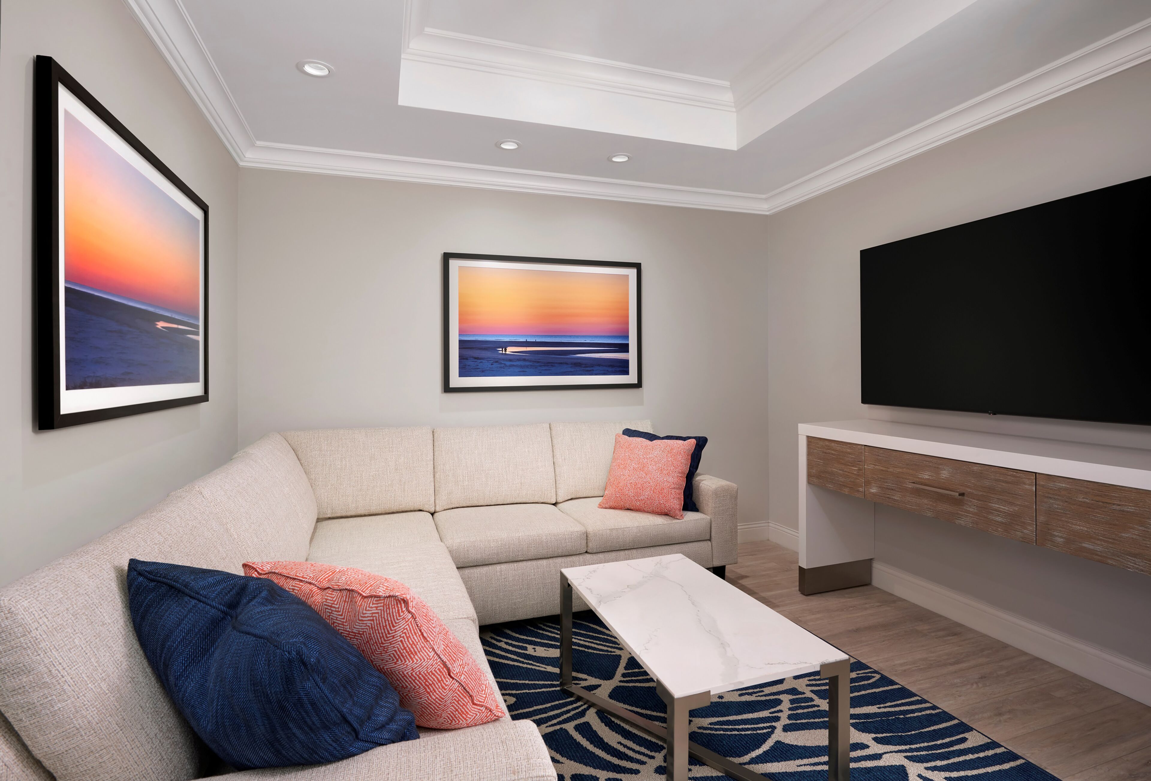 Een moderne woonkamer met een hoekbank, een flatscreen televisie en twee ingelijste zonsondergangschilderijen aan de muur.