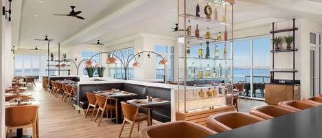 Un restaurante moderno con techos altos, varios ventiladores de techo y una zona de bar con un expositor de vidrio.