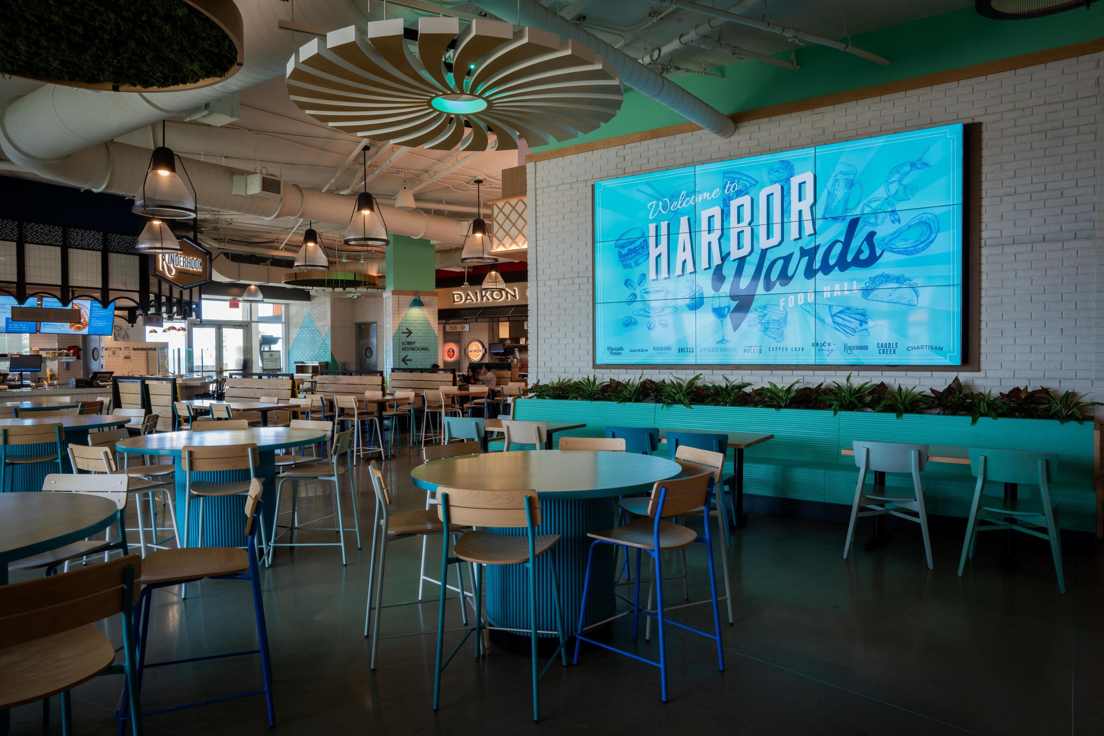 A spacious indoor dining area with multiple tables and chairs, a large illuminated welcome sign, and a brick wall.