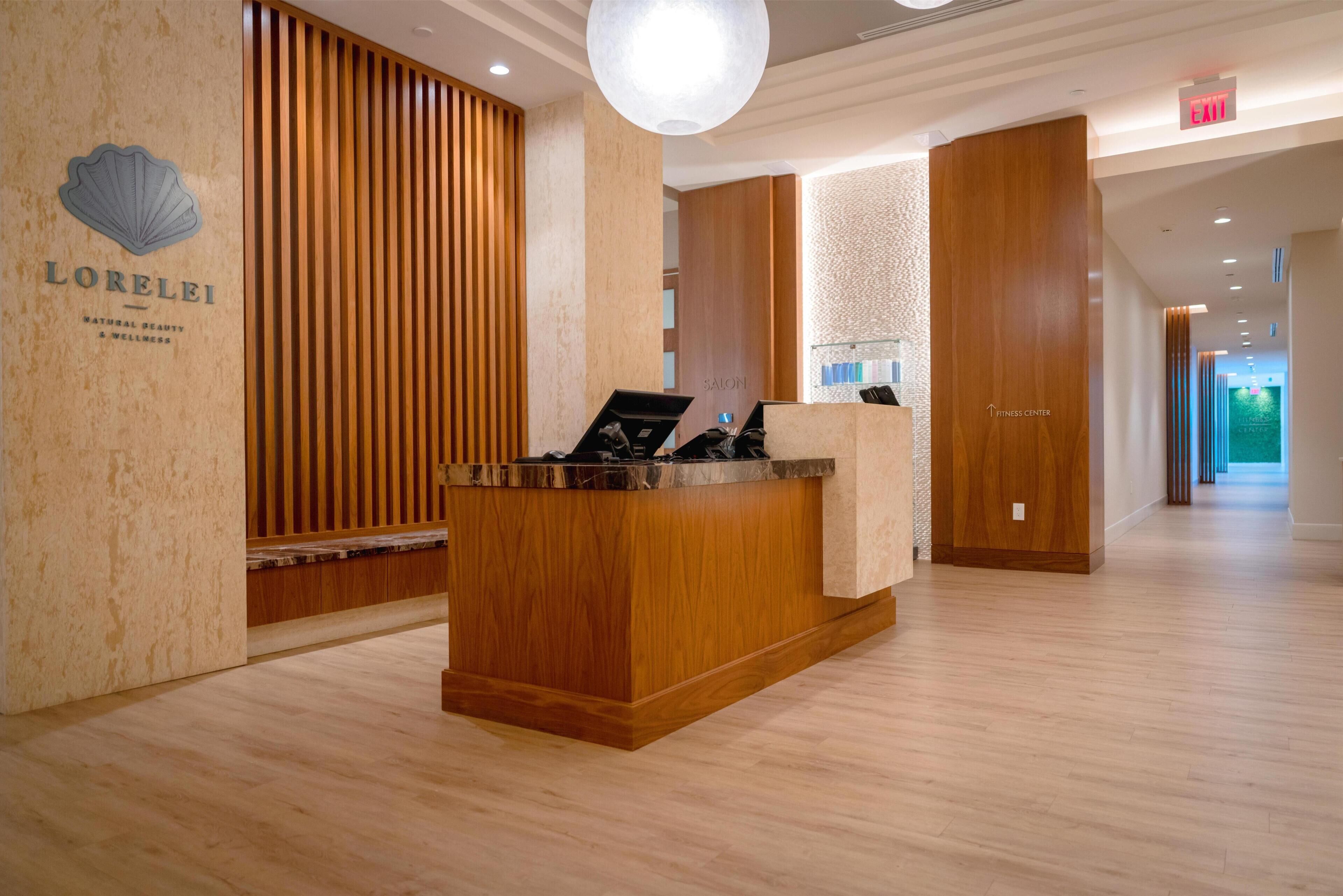 A hotel reception area with a wooden reception desk, a sign reading 'LORELEI,' and a 'Fitness Center' sign.