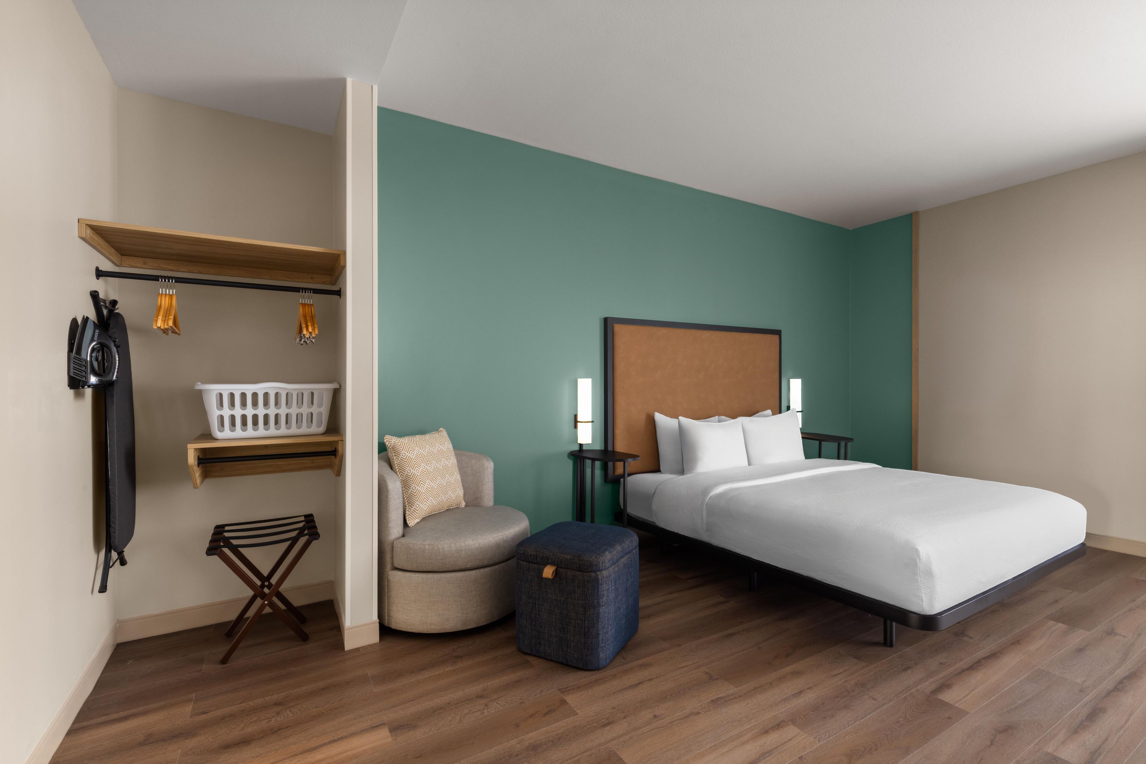 Une chambre d’hôtel avec un lit, une chaise, une table, une étagère avec des vêtements et un panier à linge.