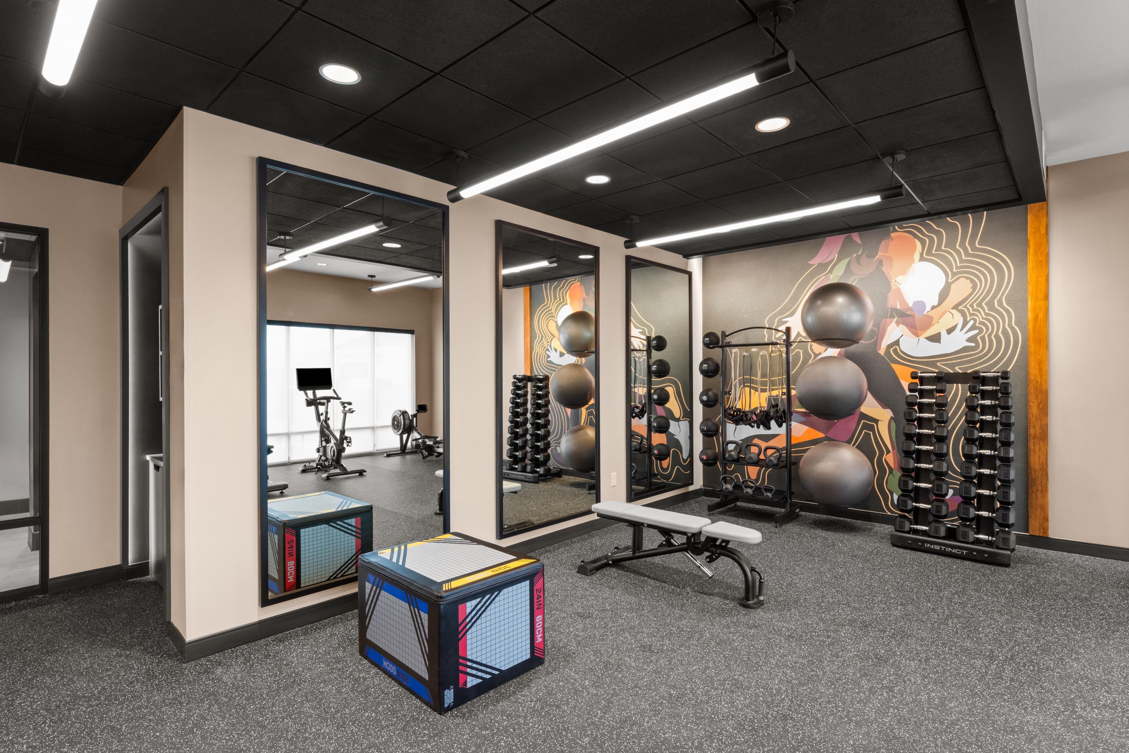 Une salle de sport équipée de matériel d’exercice, de miroirs et d’un mur orné d’une fresque colorée.