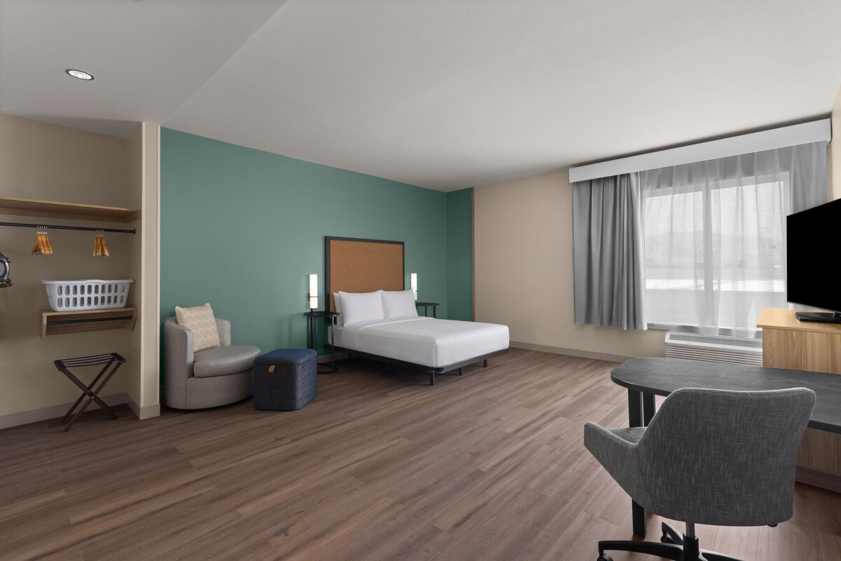 A hotel room with a bed, a desk, a chair, a small table, a wall-mounted TV, and a window with curtains.