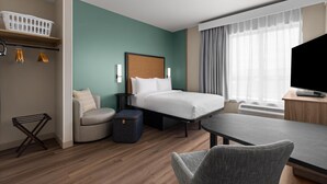 A hotel room with a bed, a desk, a chair, a TV, a window with curtains, a wall-mounted shelf, and a hanging rack with towels. - LivSmart Studios by Hilton Salt Lake City West Valley City (West Valley City)