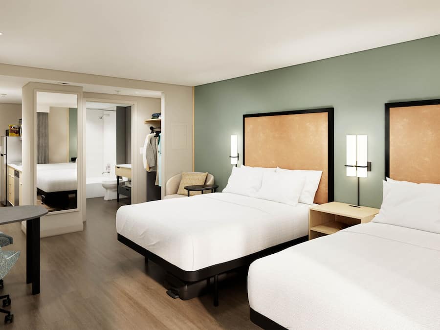 Ein Hotelzimmer mit zwei Betten, einem Schreibtisch, einem Stuhl, einem kleinen Tisch, einem Badezimmer und einer Küchenzeile.