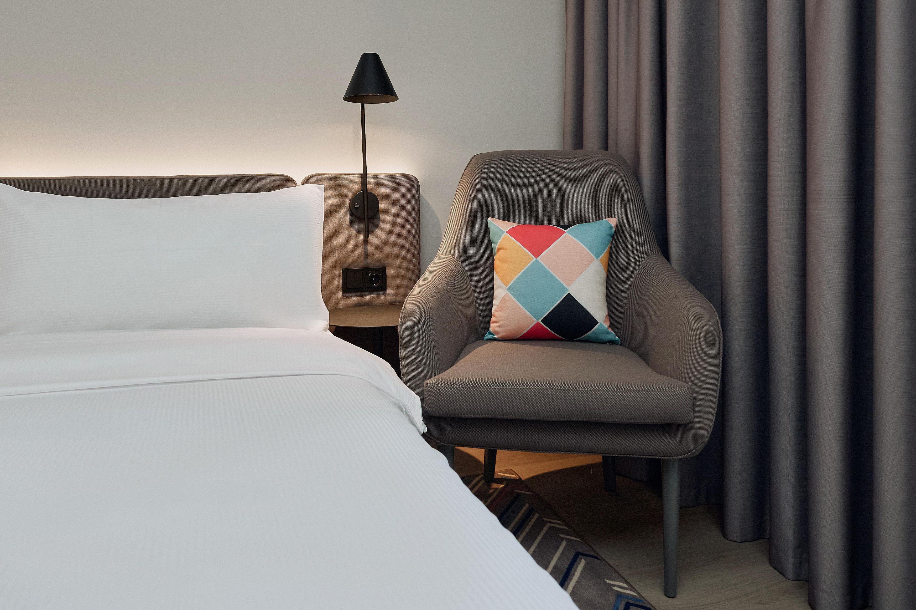 A hotel room with a bed, a chair with a patterned pillow, a nightstand with a lamp, and a curtain.