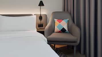A hotel room with a bed, a chair with a patterned pillow, a nightstand with a lamp, and a curtain.