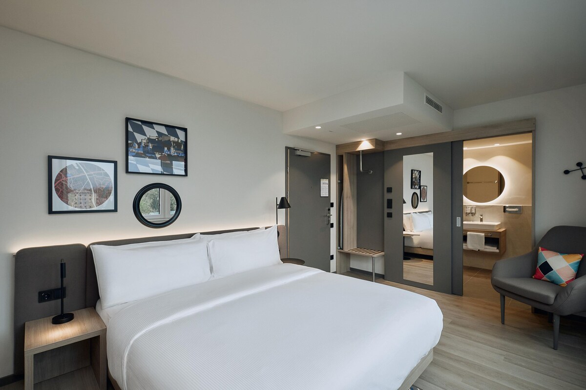 Ein modernes Hotelzimmer mit einem großen Bett, einem Schreibtisch und einem Stuhl. An der Wand befinden sich gerahmte Bilder, ein runder Spiegel und eine Tür zu einem Badezimmer.