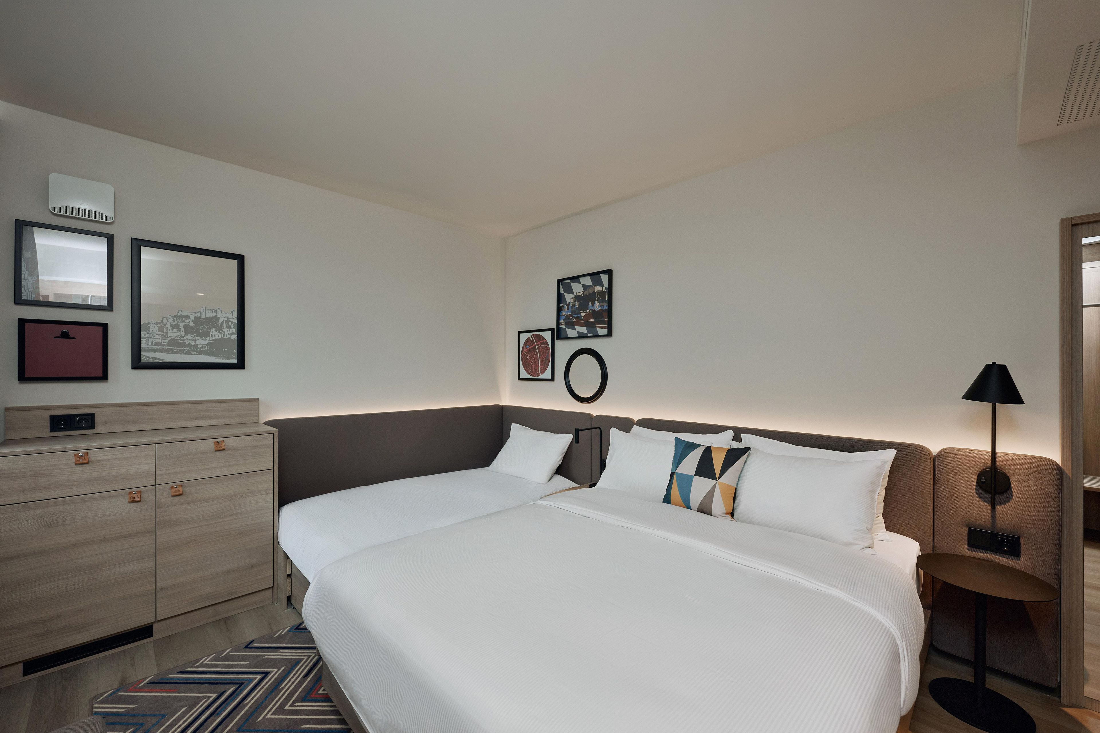 A hotel room with a large bed, a bedside table, a lamp, a dresser, and wall-mounted artwork.