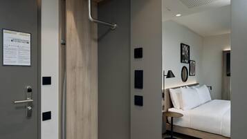 A modern hotel room with a wooden door, a bed with white bedding, a bedside table, and a wall-mounted light fixture.