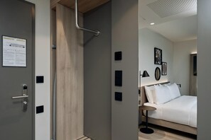 A modern hotel room with a wooden door, a bed with white bedding, a bedside table, and a wall-mounted light fixture. - Hampton By Hilton Salzburg Messe (Salzburg)