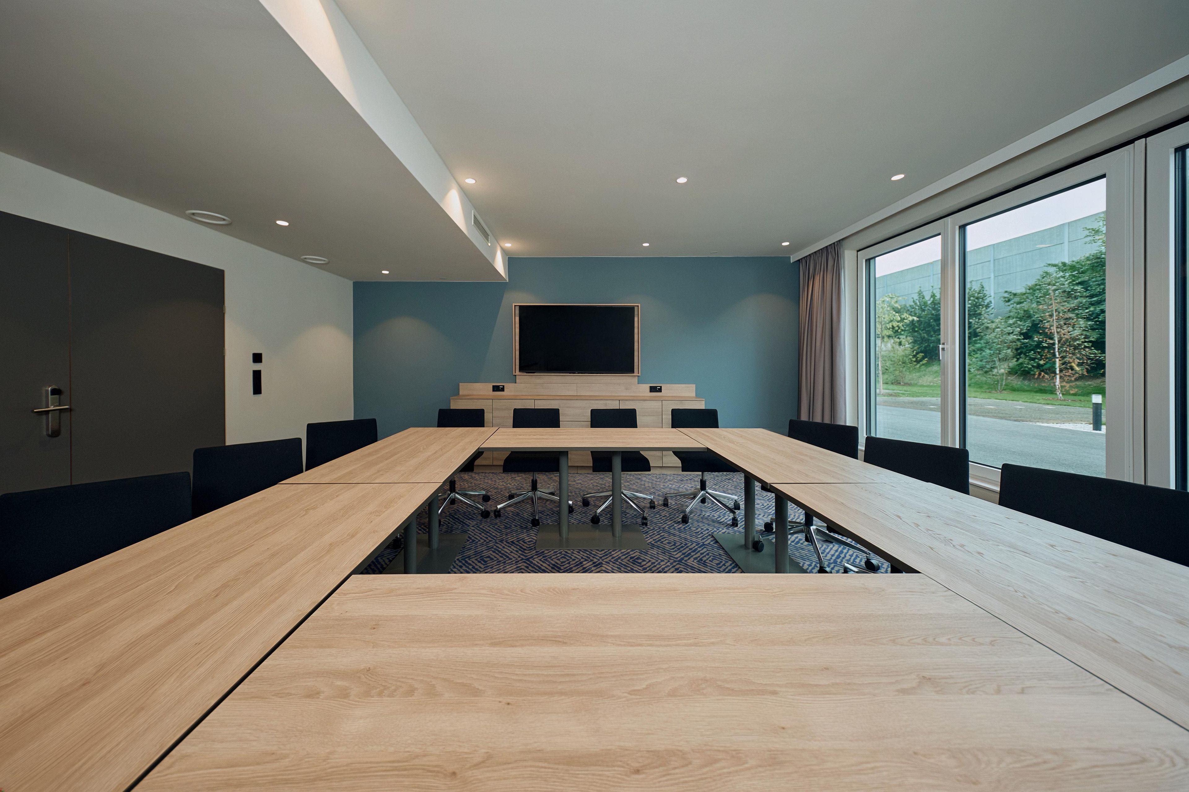 A conference room with a long table, chairs, a large screen, and a view through the windows.