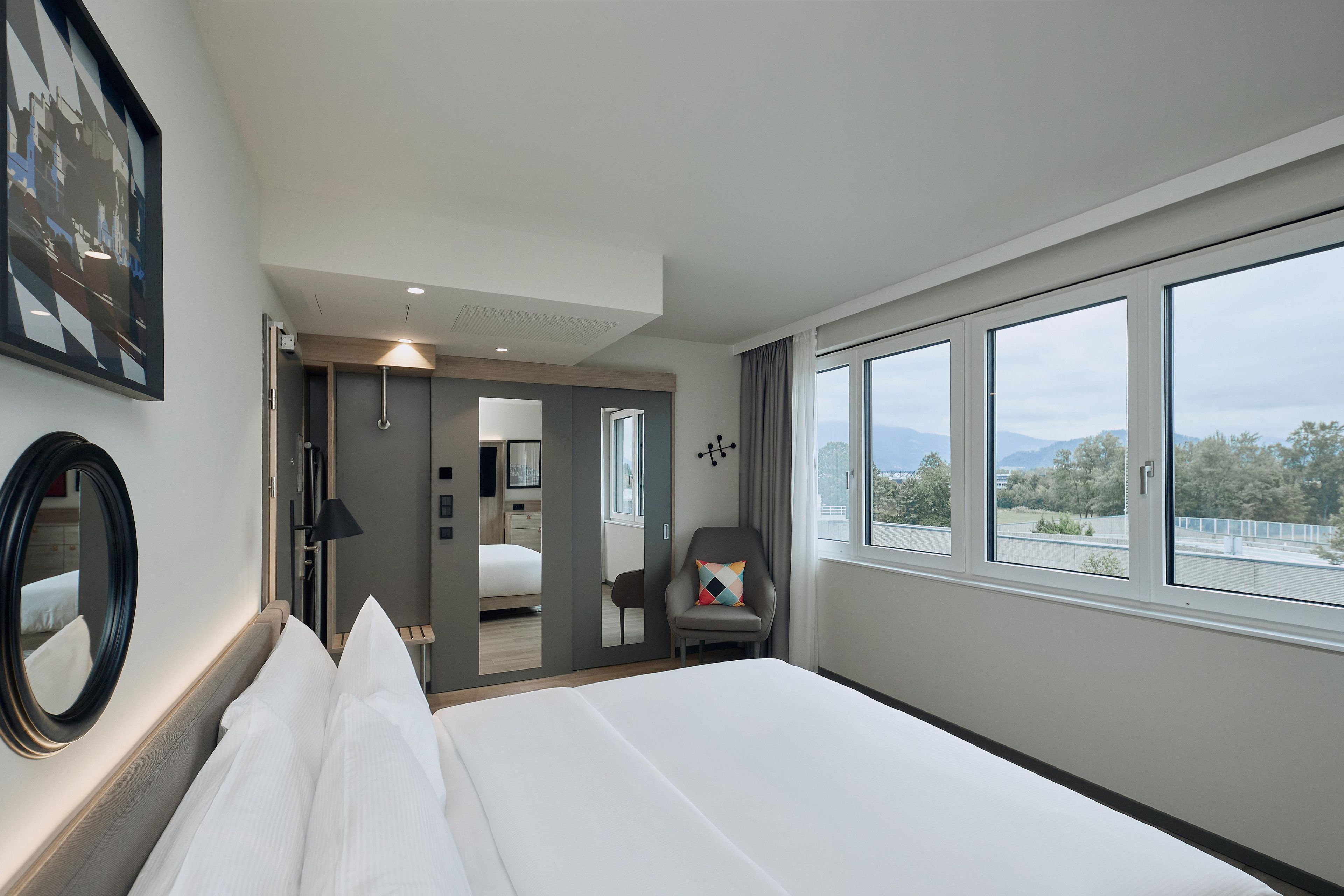 Una habitación de hotel con cama, una silla, un ventanal grande y una puerta que da a un baño.