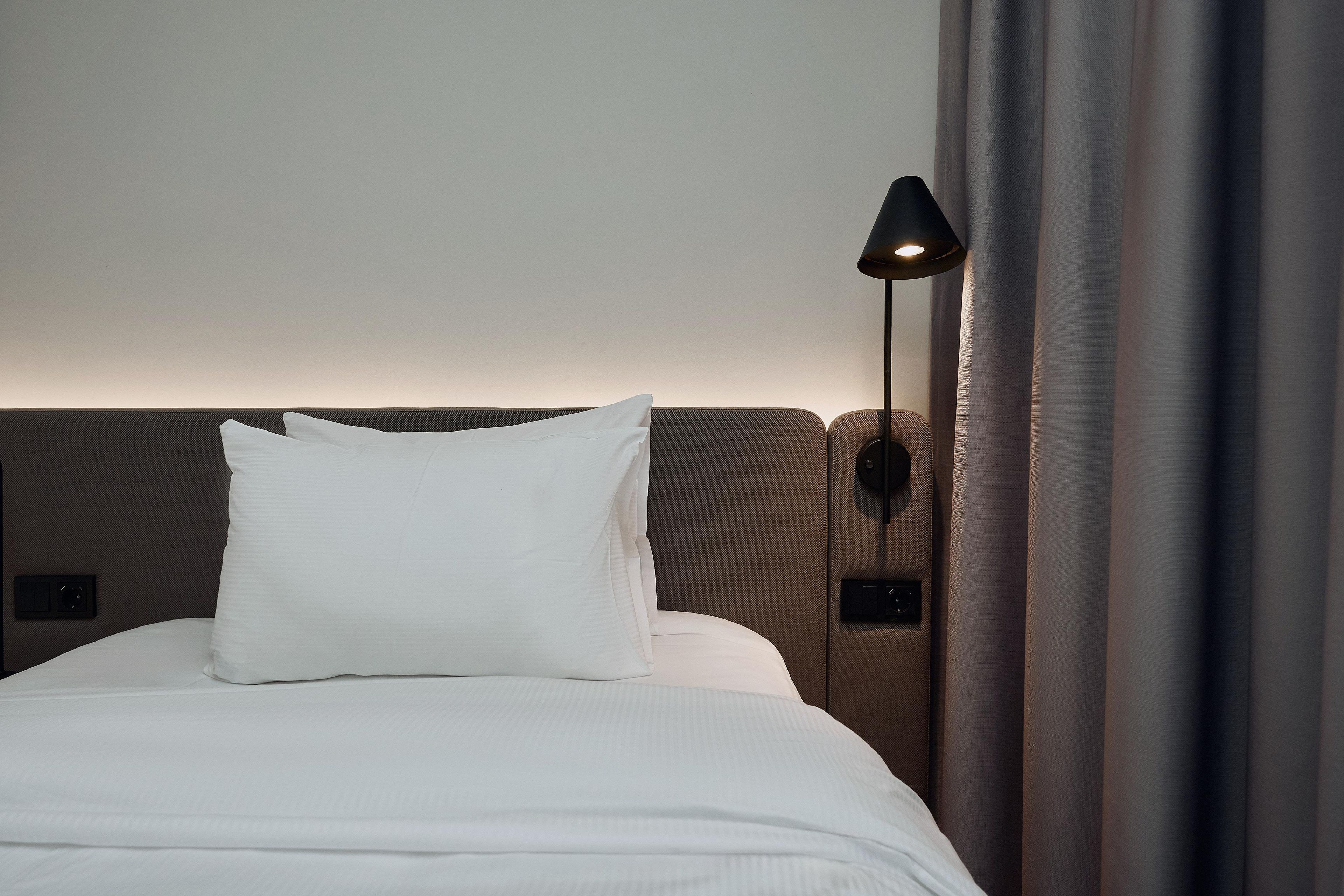 Una cama bien tendida con sábanas blancas y una única almohada blanca, una lámpara de pared y una cortina entreabierta.