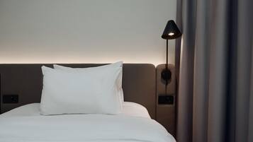 Una cama bien tendida con sábanas blancas y una única almohada blanca, una lámpara de pared y una cortina entreabierta.
