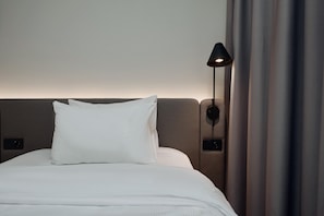A neatly made bed with white linens and a single white pillow, a wall-mounted lamp, and a curtain partially drawn to the side.