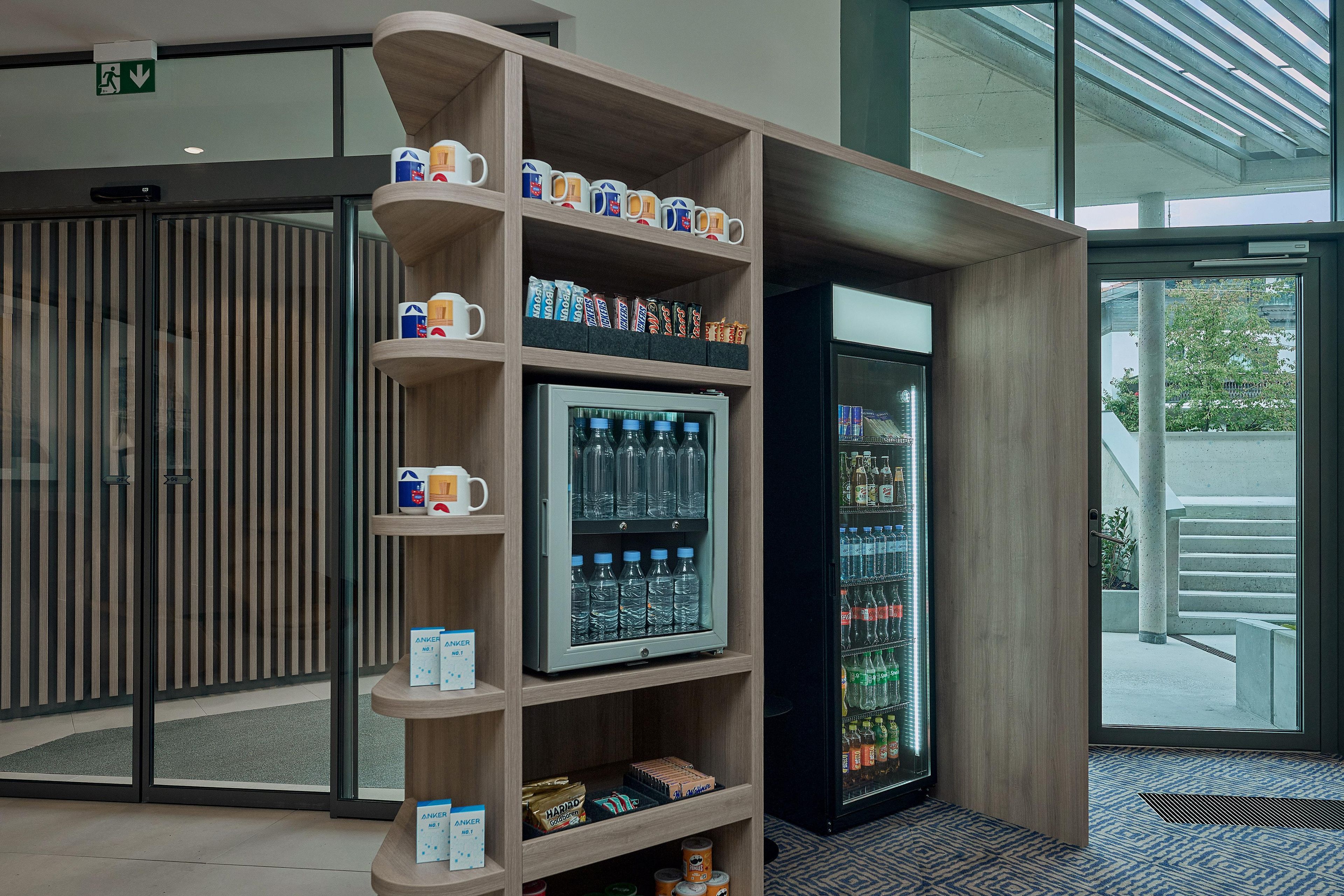 Een automaat met diverse drankjes en snacks, een koffiehoek met mokken, en een glazen deur die naar buiten leidt.