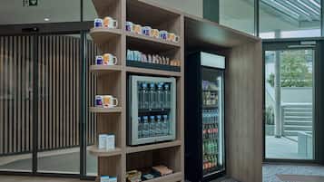 Een automaat met diverse drankjes en snacks, een koffiehoek met mokken, en een glazen deur die naar buiten leidt.