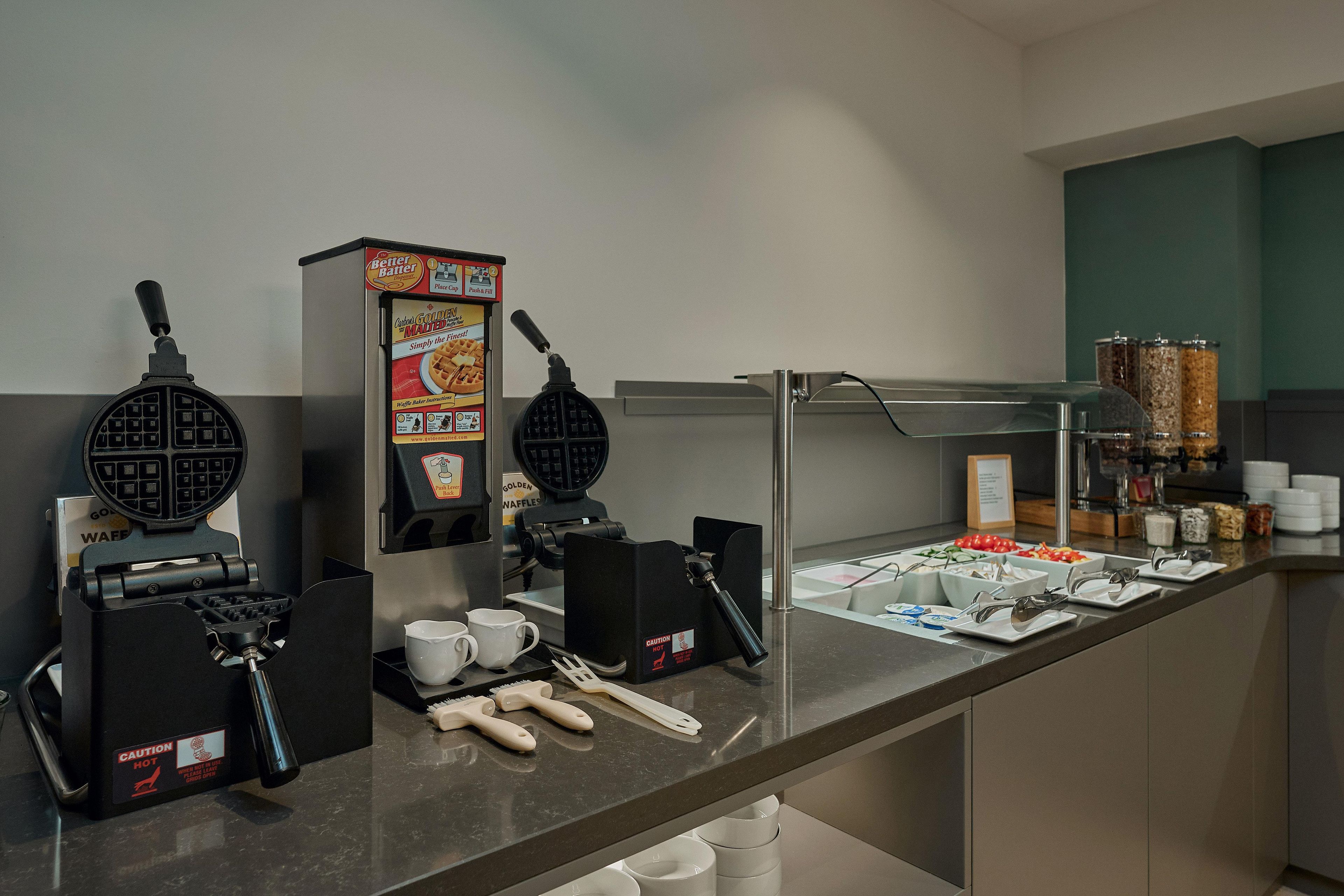A self-service breakfast bar with waffle makers, coffee machines, and a variety of food options.