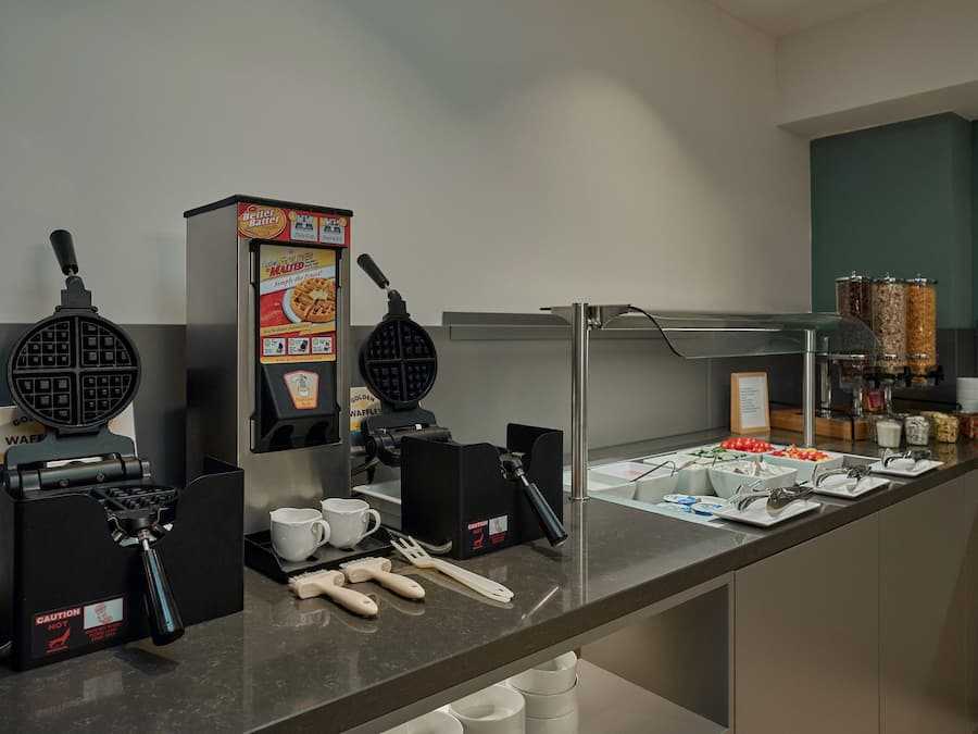 A self-service breakfast bar with waffle makers, coffee machines, and a variety of food options.