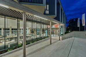 A modern building with a covered outdoor seating area, large windows, and a sign for 'Hampton by Hilton'.