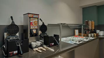 Un bufé de desayuno autoservicio con máquinas para hacer gofres, cafeteras y diversas opciones de alimentos.
