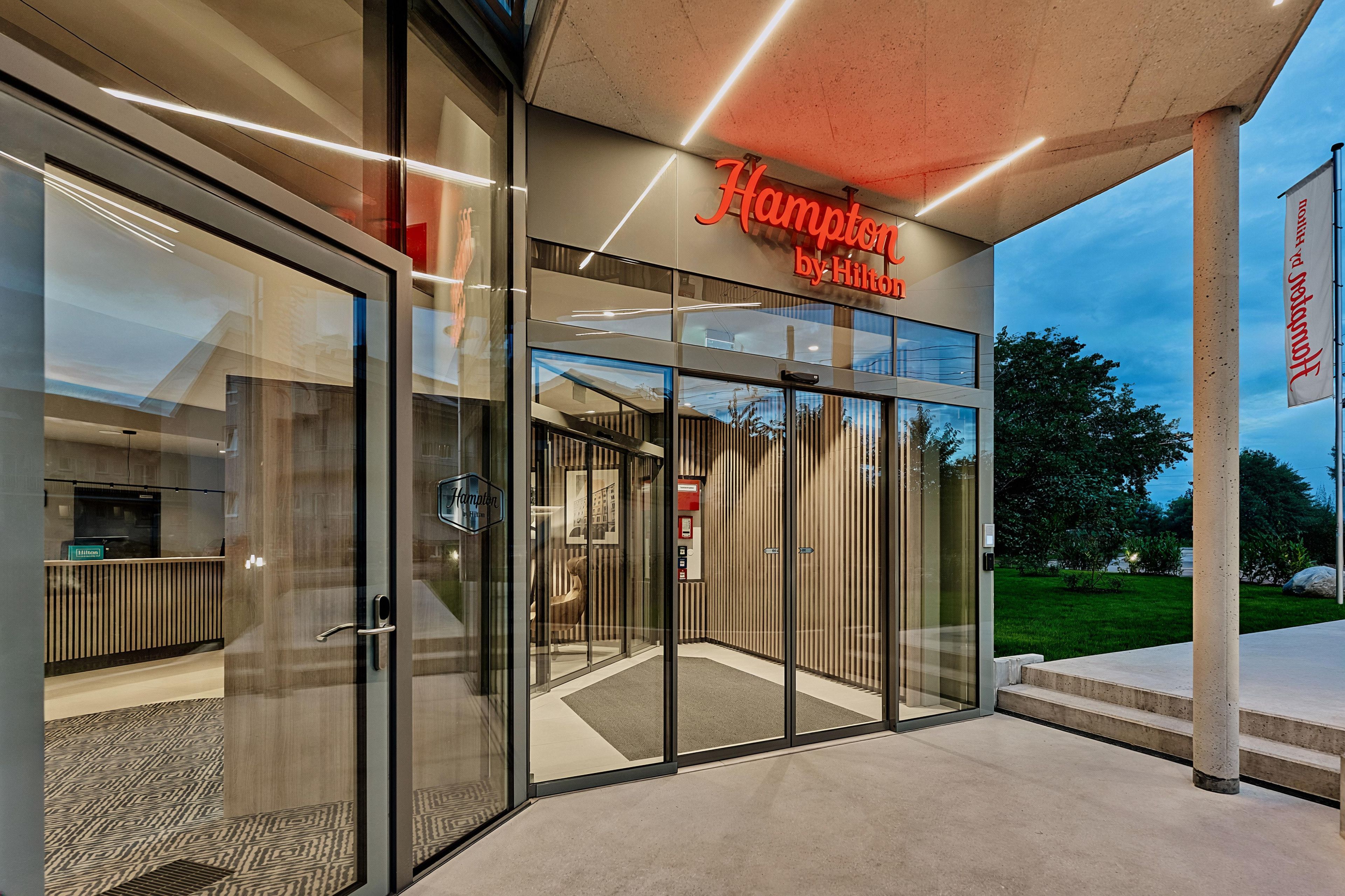 A modern hotel entrance with a glass facade, a visible elevator, and a sign indicating it's a Hampton by Hilton property.