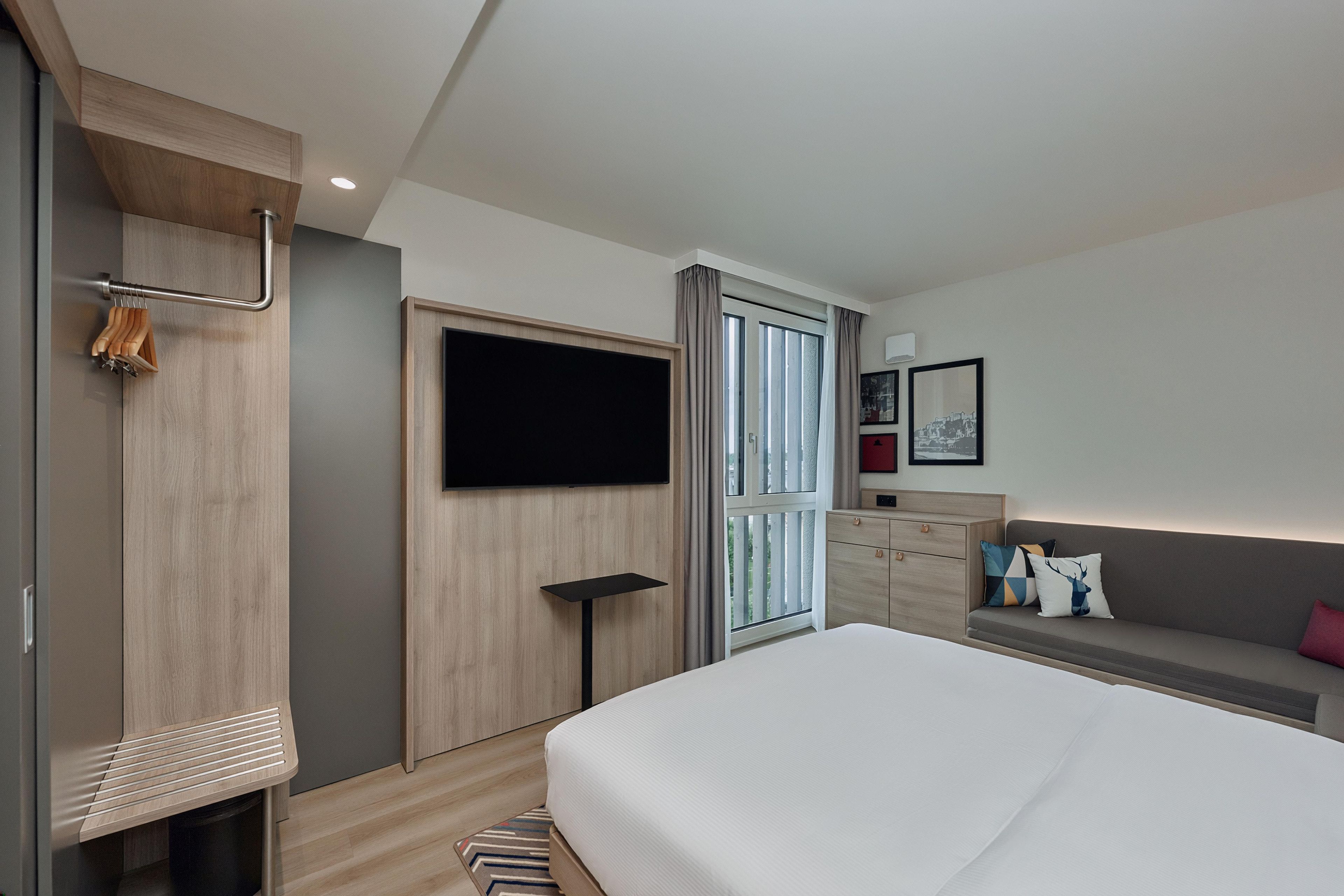 A modern hotel room with a bed, a TV, a wardrobe, and a seating area with a sofa and cushions.
