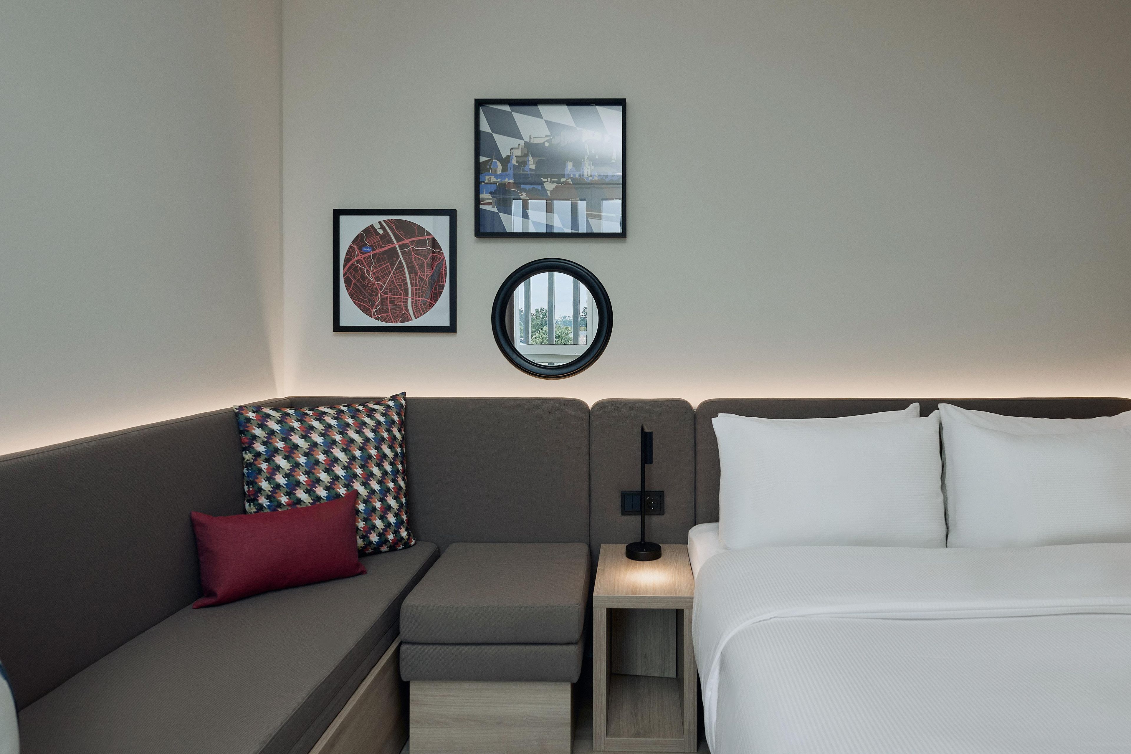 Moderni hotellihuone, jossa on harmaa sohva, pieni yöpöytä, lamppu ja valkoisilla lakanoilla varustettu sänky.
