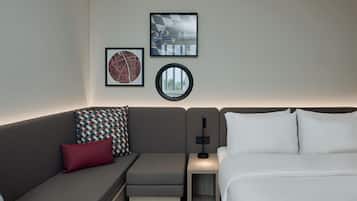 Een moderne hotelkamer met een grijze bank, een klein nachtkastje, een lamp en een bed met witte lakens.