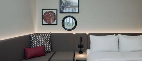 Ein modernes Hotelzimmer mit grauem Sofa, einem kleinen Nachttisch, einer Lampe und einem Bett mit weißen Leinen.