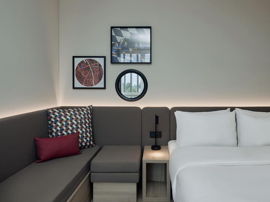 Habitación de hotel moderna con un sofá gris, una mesita de noche pequeña, una lámpara y una cama con ropa blanca.