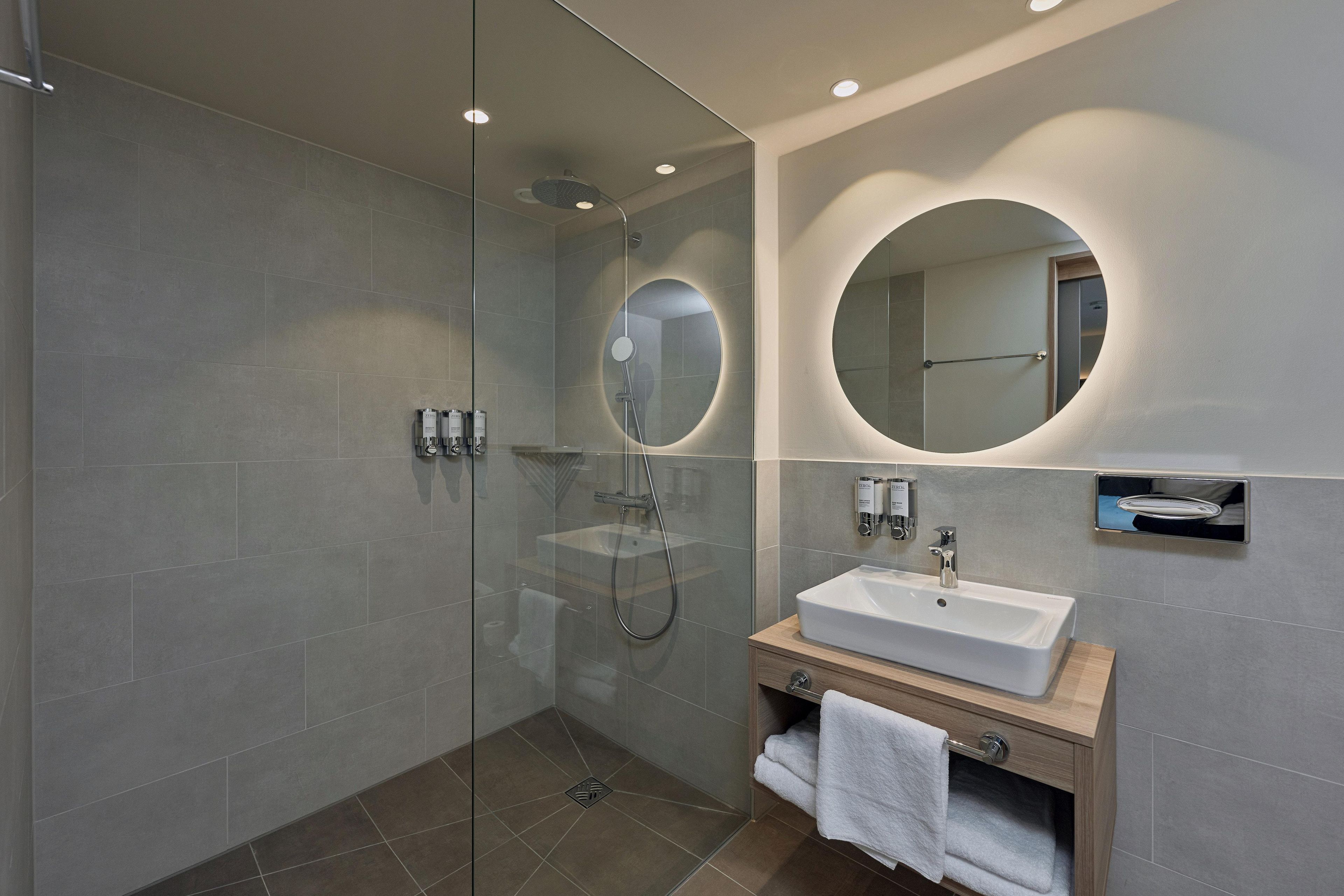 A modern bathroom with a glass-enclosed shower, a sink with a wooden vanity, and a large mirror with a circular frame.