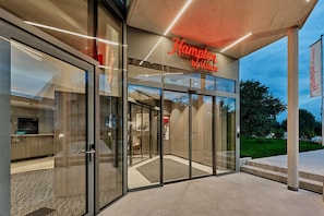 A modern hotel entrance with a glass facade, a visible elevator, and a sign indicating it's a Hampton by Hilton property. - Hampton By Hilton Salzburg Messe (Salzburg)