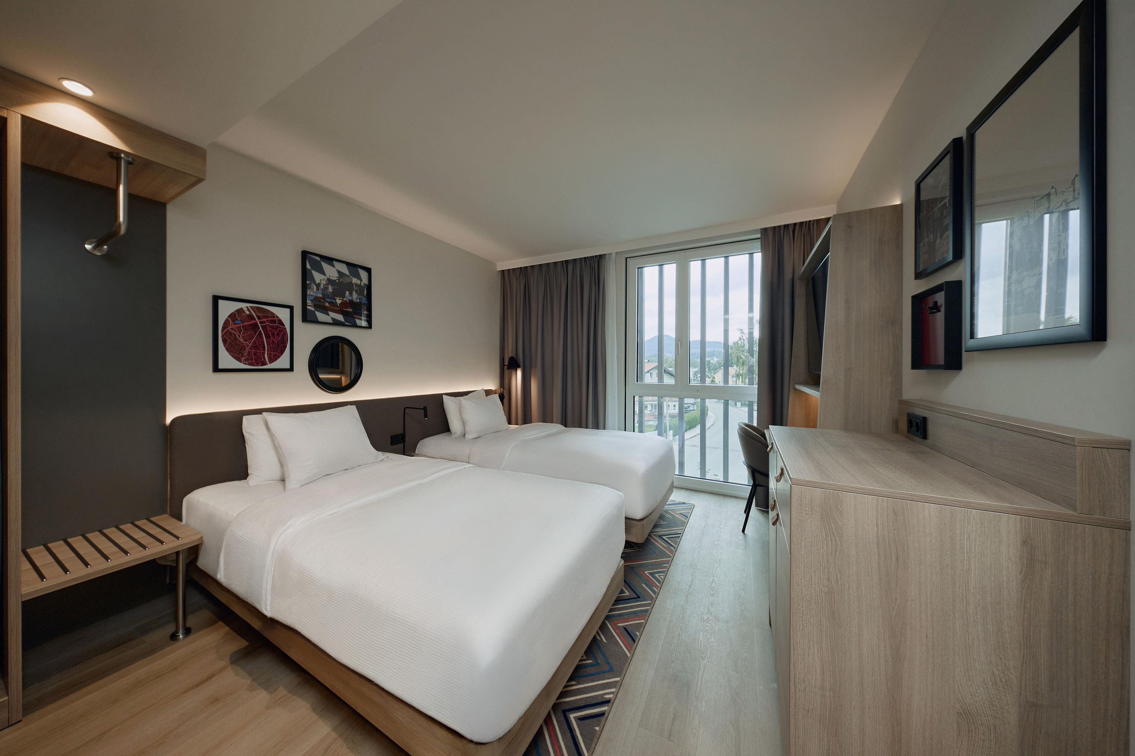 Ein Hotelzimmer mit zwei Betten, einem Schreibtisch, einem Stuhl, einem großen Fenster und Bildern an den Wänden.