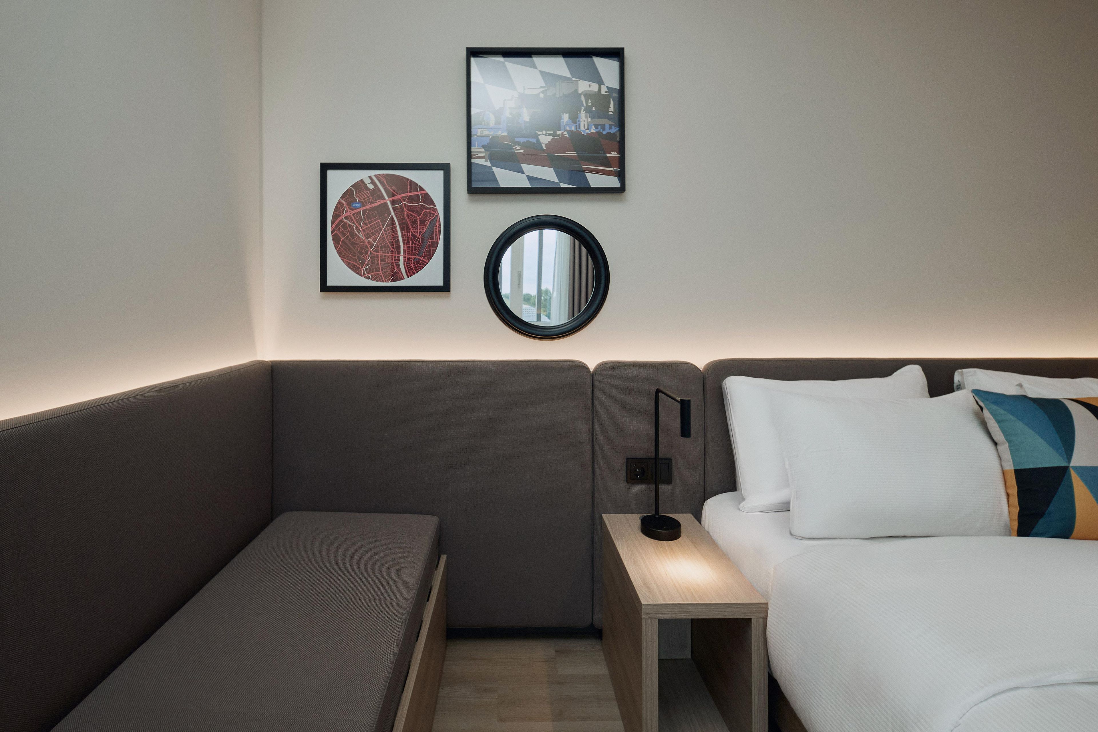 Een moderne hotelkamer met een grijze bank, een bed met witte en kleurrijke kussens, een klein nachtkastje met een lamp, en twee kunstwerken in lijsten aan de muur.
