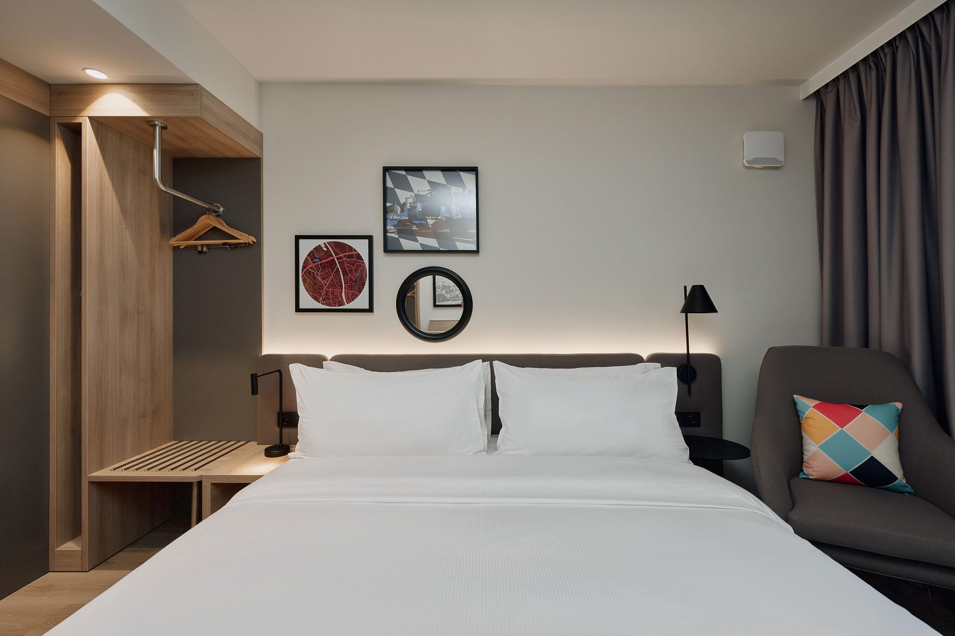 A hotel room with a bed, a bedside table, a chair, and a wall with framed pictures.