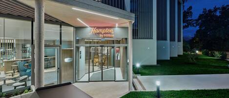 A modern building with a glass entrance, a sign reading 'Hampton by Hilton,' and a well-maintained outdoor area with grass and a pathway.