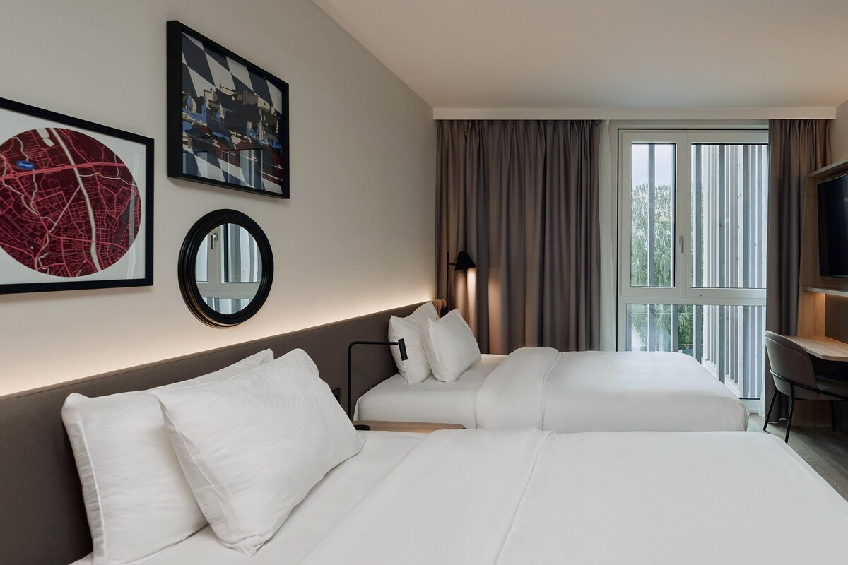 Ein Hotelzimmer mit zwei Betten, einem großen Fenster mit Vorhängen, einem Schreibtisch und einem Bild an der Wand.