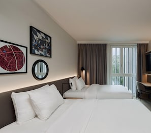 A hotel room with two beds, a large window with curtains, a desk, and artwork on the wall. - Hampton By Hilton Salzburg Messe (Salzburg)