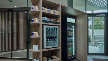 Máquina expendedora con diversas bebidas y aperitivos, estación de café con tazas y puerta de vidrio que da al exterior.