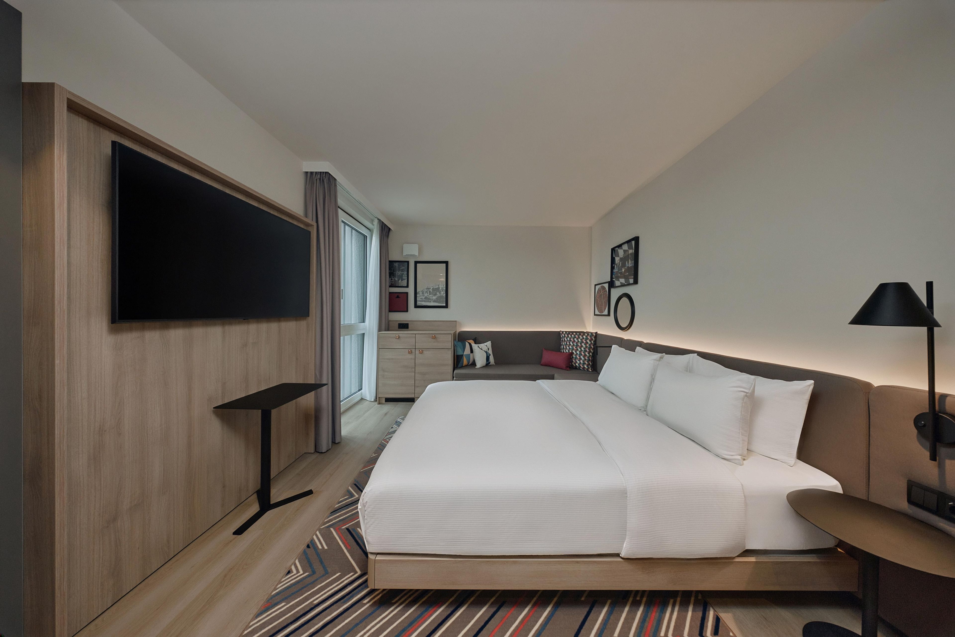 Ein Hotelzimmer mit einem großen Bett, einem an der Wand montierten Fernseher, einem Schreibtisch, einem Sessel und einem Nachttisch.