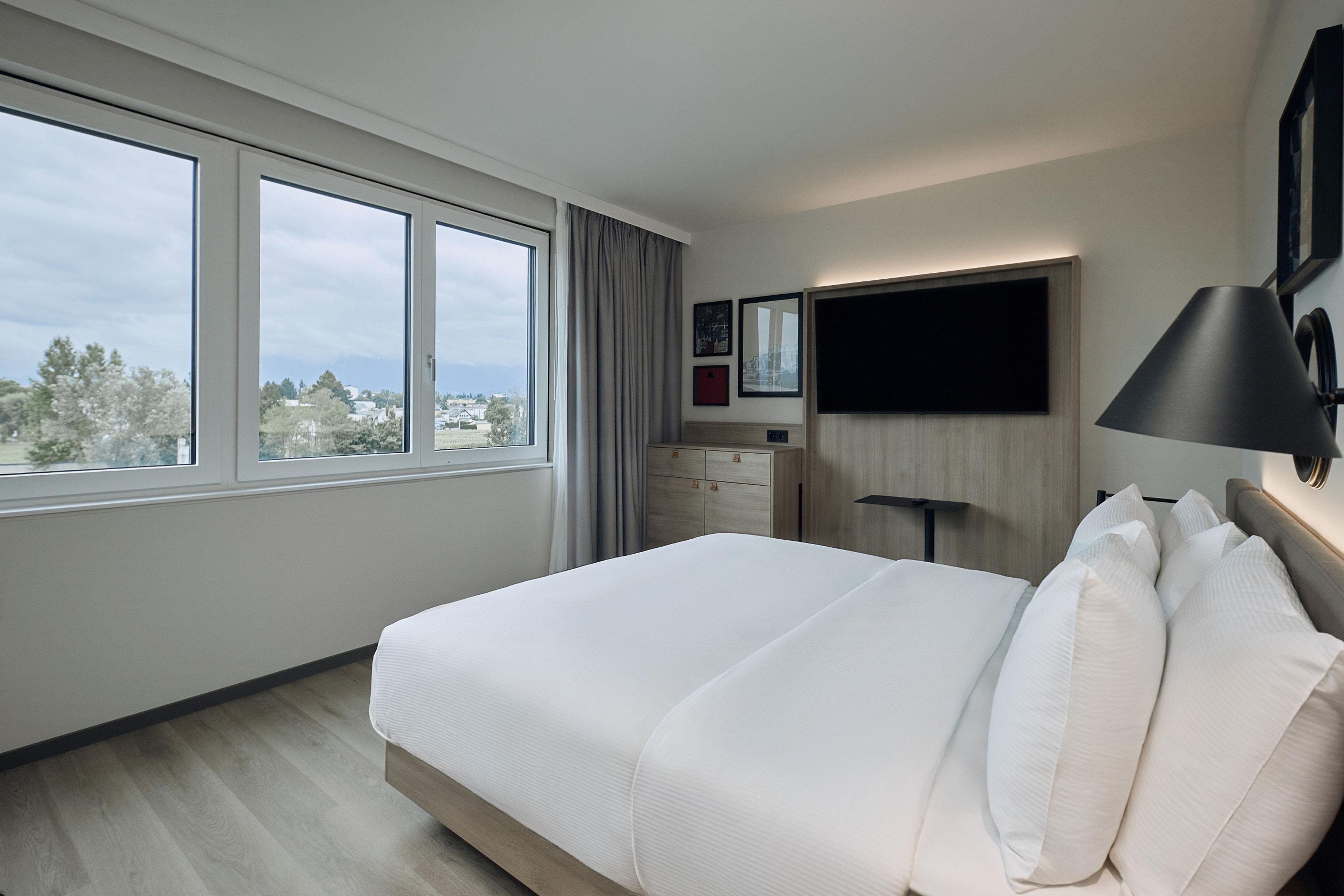 A hotel room with a large bed, a dresser, a TV mounted on the wall, and a window with a view of the city.