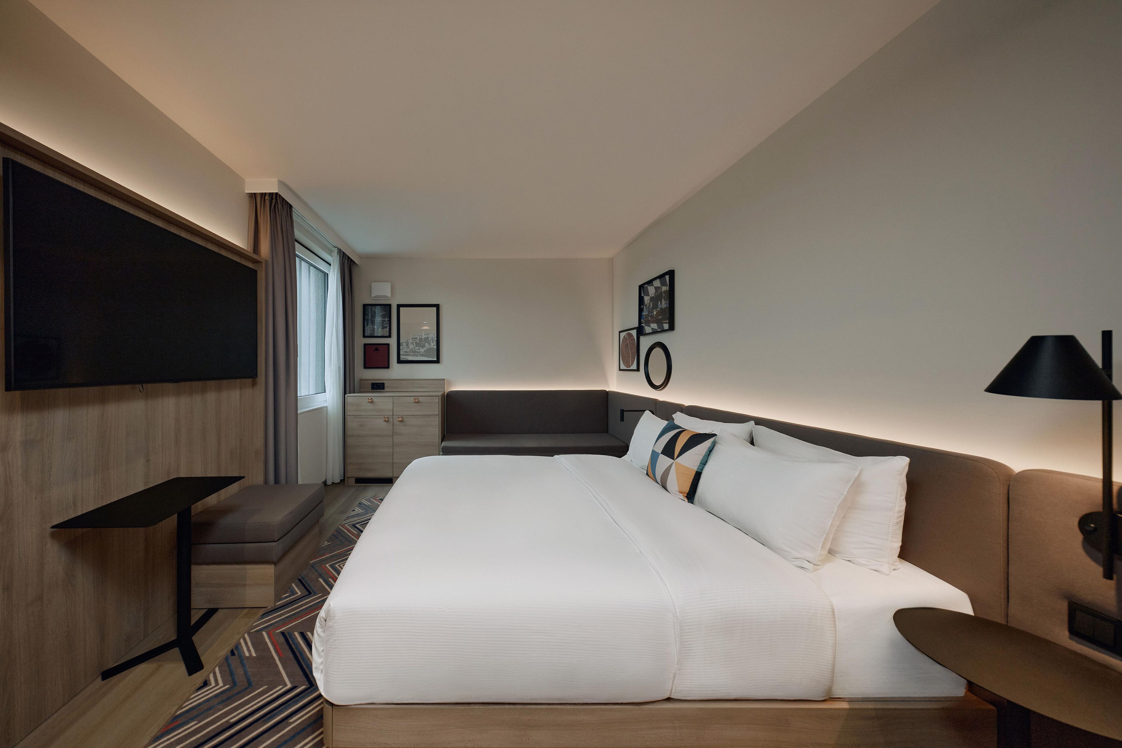 A modern hotel room with a large bed, a desk, a chair, and a TV mounted on the wall.