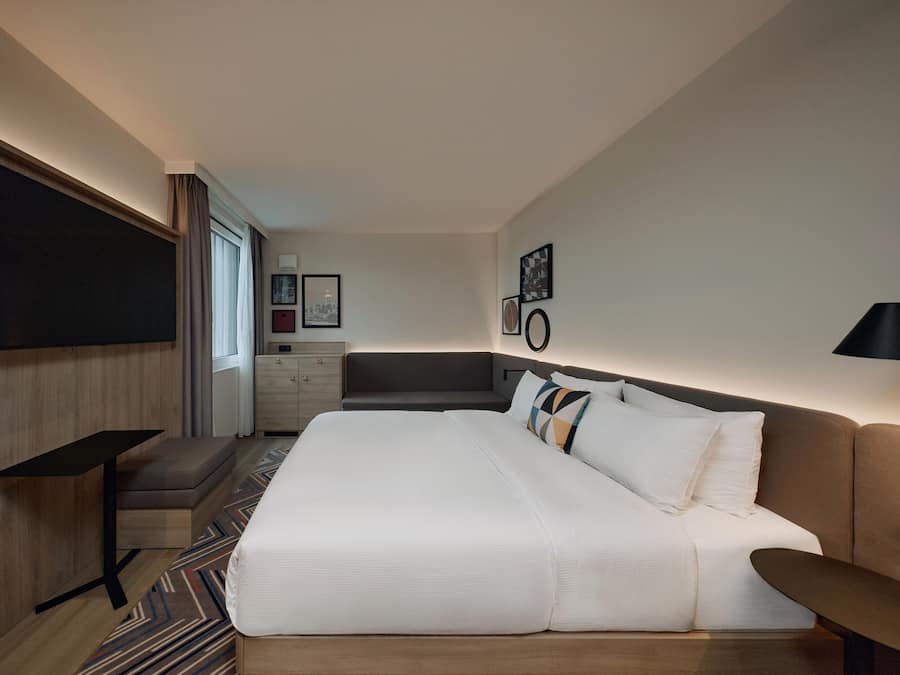 A modern hotel room with a large bed, a desk, a chair, and a TV mounted on the wall.