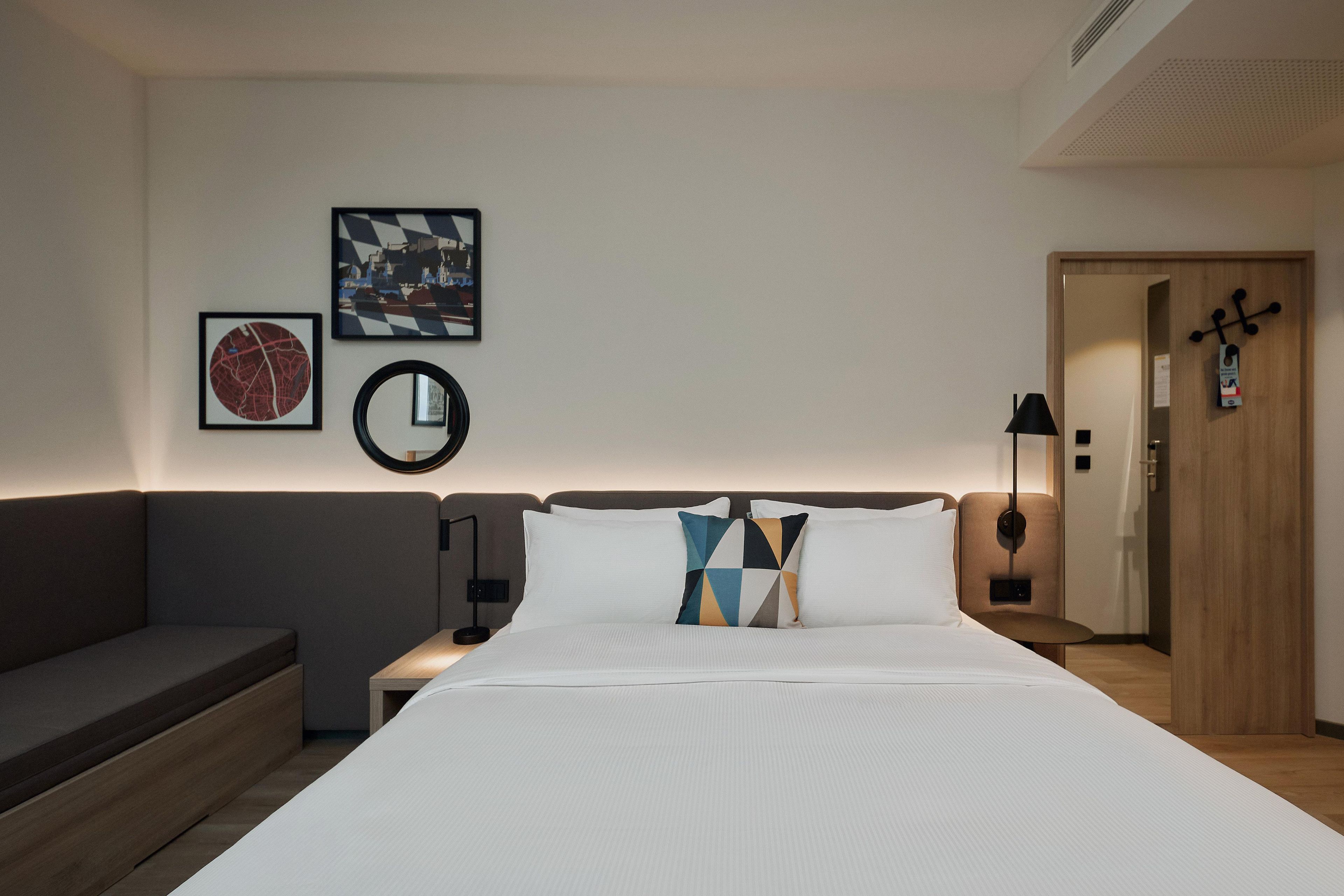 A hotel room with a bed, a nightstand, a lamp, a wall-mounted mirror, and artwork.